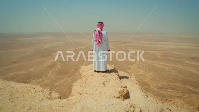 Enjoying the natural scenery, the peaceful atmosphere in the desert of the Kingdom of Saudi Arabia, a picture from behind of a Saudi Gulf Arab man wearing traditional clothing standing on top of a rocky mountain contemplating the barren desert area