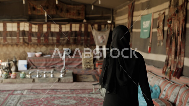 A trip and a camping trip in the winter weather in the desert, a popular traditional Bedouin tent in Saudi Arabia, the calm atmosphere in the camping places, a picture from behind of a veiled Saudi Gulf Arab woman wearing a black abaya and standing inside a verse of poetry