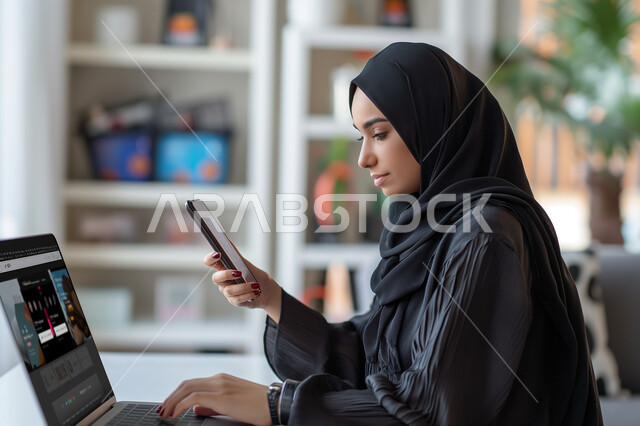 العمل ضمن مقر الشركة، إدارة شؤون الأعمال وتنظيمها, التفكير في الاستراتيجيات والأهداف ، موظفة عربية سعودية خليجية ترتدي الحجاب تحمل بيدها هاتف محمول، استخدام جهاز اللابتوب المحمول لإنجاز الأعمال داخل المكتب، إيماءات التركيز والتمعن