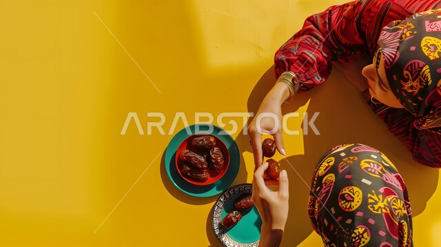 مأكولات ذات اهمية غذائية عالية, صورة من الاعلى لسيدتان عربيتان خليجيتان محجبتان يجلسان على طاولة صفراء يقومون بتناول بعض من حبات التمر الطازجه, مفهوم الكرم في الضيافة والاستقبال, خلفية ملونة