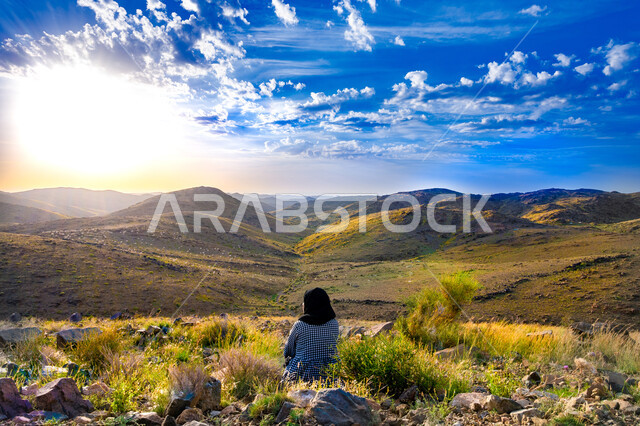 التنزه في إحدى الحدائق الترفيهية بالمملكة العربية السعودية في النهار، صورة من الخلف لفتاة عربية سعودية خليجية تجلس على الاراضي العشبية تراقب شروق الشمس، خلفية المناظر الطبيعية 