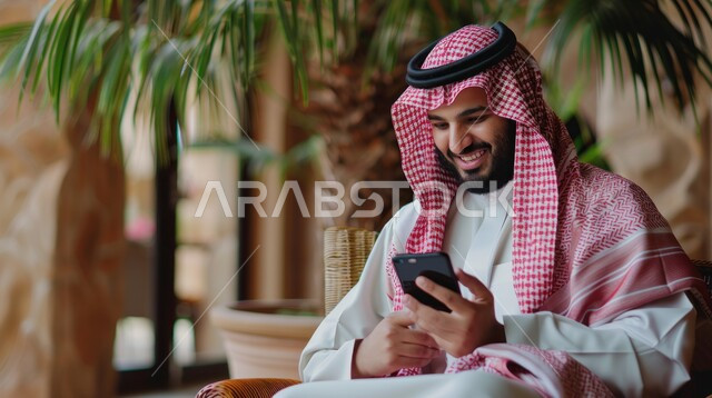 استخدام الأجهزة والتقنيات الحديثة لتسهيل الأعمال, رجل أعمال عربي سعودي خليجي يرتدي الشماغ والثوب التقليدي يجلس في مكتبه ويستخدم جهاز التابلت بتعابير السرور, إجراء مقابلات عمل أونلاين, مهن ووظائف سعودية مكتبية, التواصل مع الاهل و الاقارب