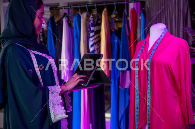 شابة عربية سعودية خليجية جميلة محجبة تبتسم أثناء عملها في مكتبها , تقوم بإبتكارموديلات رائعة , مصممة أزياء خليجية جذابة ترسم رسمًا باستخدام كمبيوتر محمول , مصممة أزياء مبتكرة , ورشة عمل تصميم ازياء , معدات الخياطة , تصميم ورسم الأزياء عن طريق اللابتوب