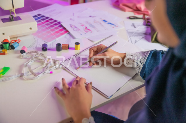 صورة مقربة من الأعلى , ليد مصممة أزياء خليجية سعودية  تعمل على تصميم ورسم الأزياء على دفتر الرسم أثناء عملها في مكتبها , مصممة أزياء مبتكرة , ورشة عمل تصميم ازياء ,  أدوات الرسم والخياطة