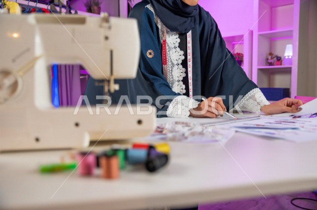 شابة عربية سعودية خليجية جميلة محجبة تبتسم أثناء عملها في مكتبها , تقوم بإبتكارموديلات رائعة , مصممة أزياء خليجية تعمل على تصميم ورسم الأزياء على ورق ابيض , مصممة أزياء مبتكرة , ورشة عمل تصميم ازياء
