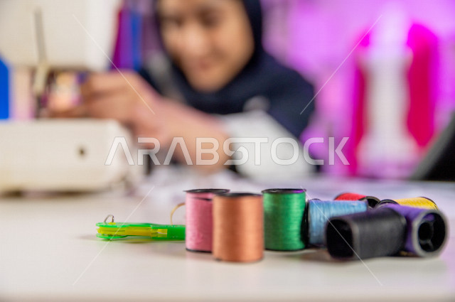 شابة عربية سعودية خليجية جميلة محجبة تبتسم أثناء عملها في مكتبها , مصممة أزياء خليجية جذابة تخيط الملابس بماكينة الخياطة , مصممة أزياء مبتكرة , ورشة عمل تصميم ازياء , معدات وآلة الخياطة , تصميم وخياطة الملابس