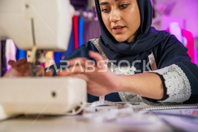 شابة عربية سعودية خليجية جميلة محجبة تبتسم أثناء عملها في مكتبها , مصممة أزياء خليجية جذابة تخيط الملابس بماكينة الخياطة , مصممة أزياء مبتكرة , ورشة عمل تصميم ازياء , معدات وآلة الخياطة , تصميم وخياطة الملابس