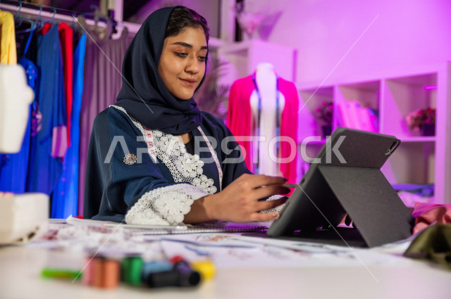 A beautiful veiled Saudi Arabian Gulf young woman smiling while working in her office, creating amazing models, an attractive Gulf fashion designer who draws a drawing using a laptop, an innovative fashion designer, a fashion design workshop, sewing equip