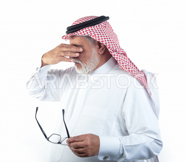 بورتريه رجل عربي سعودي كبير بالسن يشعر بالإجهاد النفسي والصداع بعد إرتداء النظارة , يرتدي ثوب أبيض مع شماغ وعقال , خلفية بيضاء , رجل مسن متعب يشعر بألم في مقدمة رأسه , مفهوم لغة الجسد وتعابيرالوجه