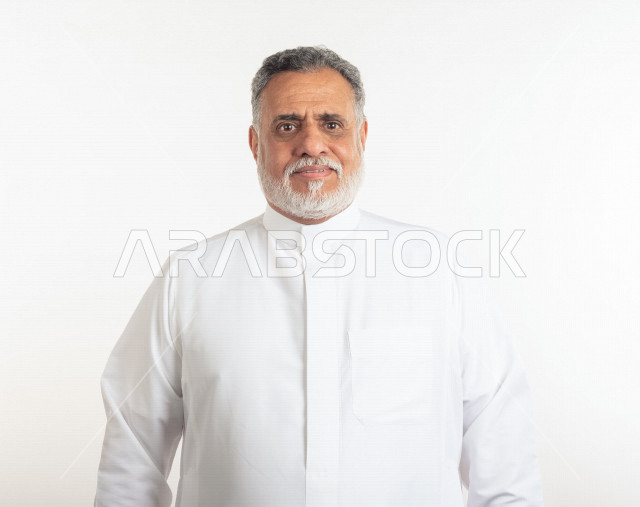 بورتريه رجل عربي سعودي كبير بالسن مبتسم يقوم بجلسة تصوير , يرتدي ثوب أبيض , خلفية بيضاء , رجل مسن بشوش الوجه يلتقط الصور , مفهوم تعابيرالوجه ولغة الجسد