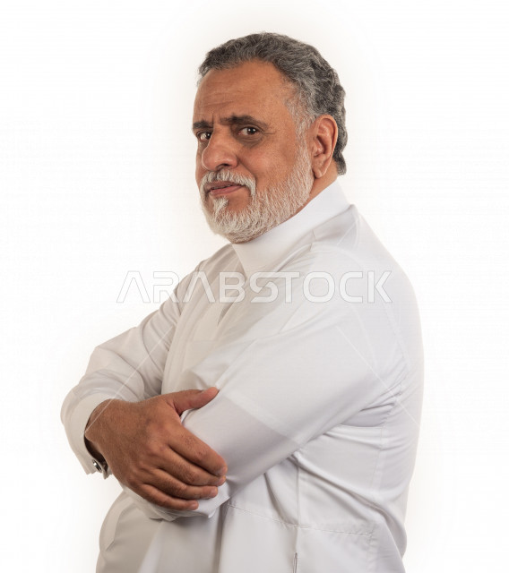 بورتريه رجل عربي سعودي كبير بالسن مبتسم يقوم بجلسات التصوير بأيدي متقاطعة , يرتدي ثوب أبيض , خلفية بيضاء , منظر جانبي لرجل مسن بشوش الوجه يلتقط الصور , مفهوم تعابيرالوجه ولغة الجسد