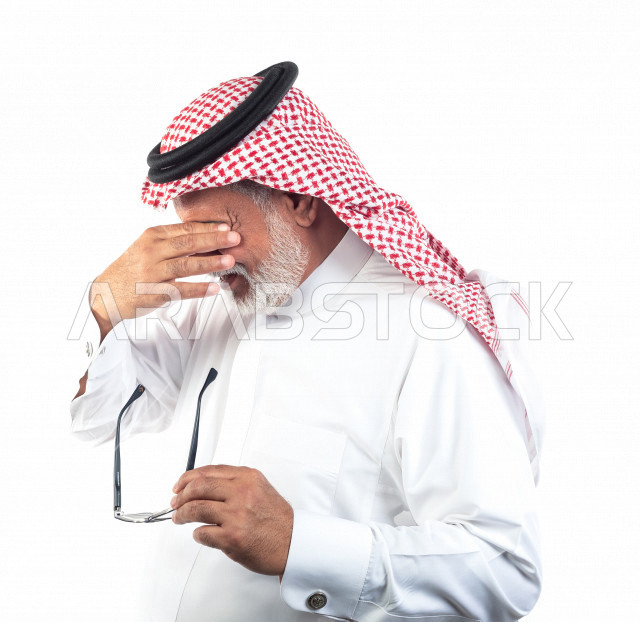 Portrait of an old Saudi Arab man feeling psychological stress and headache after wearing glasses, wearing a white dress with a shemagh and headband, white background, a tired old man feeling pain in the front of his head, the concept of body language and