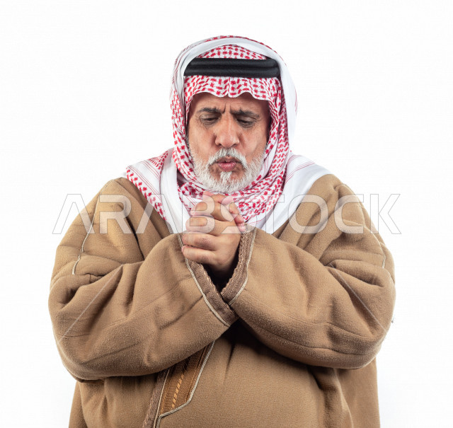 بورتريه رجل عربي سعودي كبير بالسن يشعر بالبرد , يرتدي البشت السعودي مع شماغ وعقال , خلفية بيضاء , رجل مسن يشعر بحالة من البرد والصقيع مفهوم لغة الجسد وتعابيرالوجه المختلفة