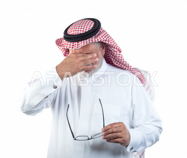 بورتريه رجل عربي سعودي كبير بالسن يشعر بالإجهاد النفسي والصداع بعد إرتداء النظارة , يرتدي ثوب أبيض مع شماغ وعقال , خلفية بيضاء , رجل مسن متعب يشعر بألم في مقدمة رأسه , مفهوم لغة الجسد وتعابيرالوجه