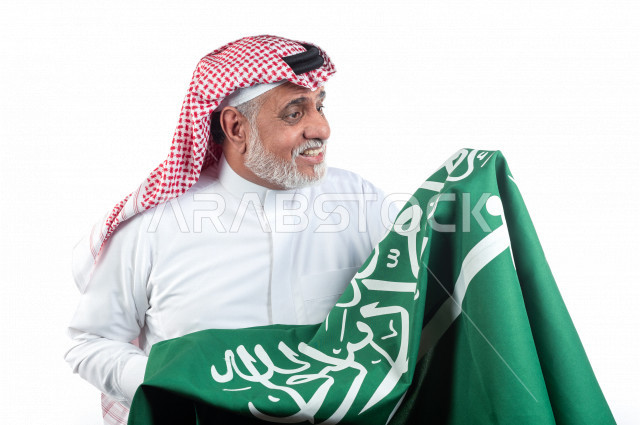 بورتريه رجل عربي سعودي كبير بالسن يحمل علم المملكة العربية السعودية وحالة من السعادة , يرتدي ثوب أبيض مع شماغ وعقال , خلفية بيضاء , رجل مسن متبسم وهو يحمل علم السعودية مفهوم تعبيرالوجه 