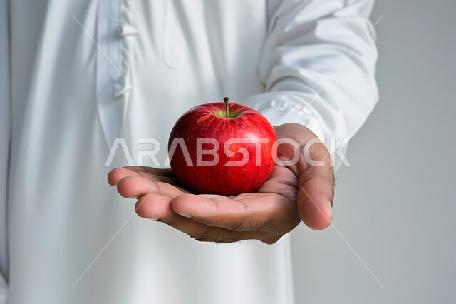 انتقاء أجود أنواع الفواكه الطازجة، المحاصيل الزراعية السعودية ذات الجودة العالية، بورتريه مقرب لشاب عربي خليجي سعودي يرتدي الثوب التقليدي يحمل بيده تفاحة حمراء، وجبة طعام صحية خفيفة، تلبية الحاجات الضرورية للجسم، خلفية بيضاء