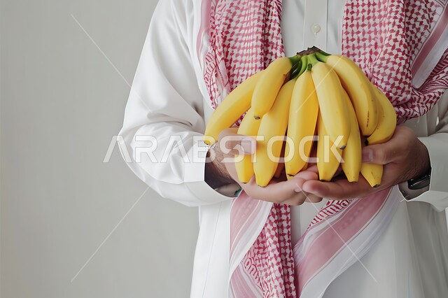 تلبية الحاجات الضرورية للجسم، وجبة طعام صحية خفيفة، اختيار أجود أنواع الفواكه الطازجة، المحاصيل الزراعية السعودية ذات الجودة العالية، بورتريه مقرب لشاب عربي خليجي سعودي يرتدي الثوب التقليدي والشماغ يحمل بيده حبات الموز، خلفية بيضاء