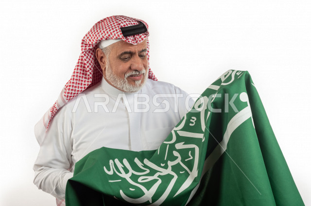 بورتريه رجل عربي سعودي كبير بالسن يحمل علم المملكة العربية السعودية وحالة من السعادة , يرتدي ثوب أبيض مع شماغ وعقال , خلفية بيضاء , رجل مسن متبسم وهو يحمل علم السعودية مفهوم تعبيرالوجه 
