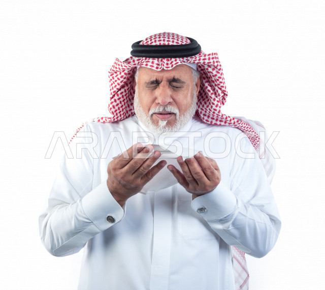 بورتريه رجل عربي سعودي كبير بالسن بيده منديل يضعه على أنفه والرغبة بالعطس , رجل شايب سعودي يرتدي ثوب أبيض مع شماغ وعقال , خلفية بيضاء , رجل مسن في حاله من العطس مفهوم لغة الجسد وتعابير الوجه
