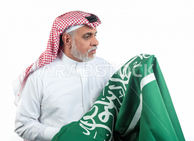 بورتريه رجل عربي سعودي كبير بالسن يحمل علم المملكة العربية السعودية وحالة من السعادة , يرتدي ثوب أبيض مع شماغ وعقال , خلفية بيضاء , رجل مسن متبسم وهو يحمل علم السعودية مفهوم تعبيرالوجه 