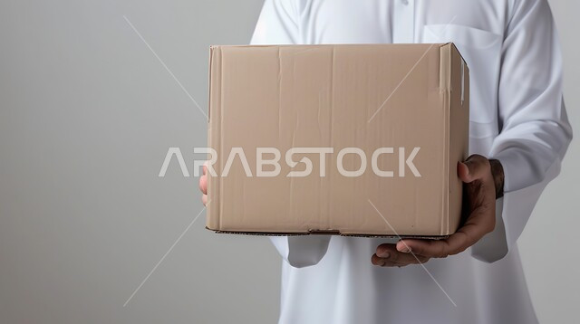 عملية توصيل الطلبات للزبائن، الشحن والتوصيل ذات الجودة العالية، بورتريه مقرب لرجل عربي خليجي سعودي يرتدي الزي التقليدي يعمل كمندوب مبيعات ويحمل بيده صندوق كرتوني، التسوق وطلب المنتجات عبر الإنترنت، خلفية رمادية
