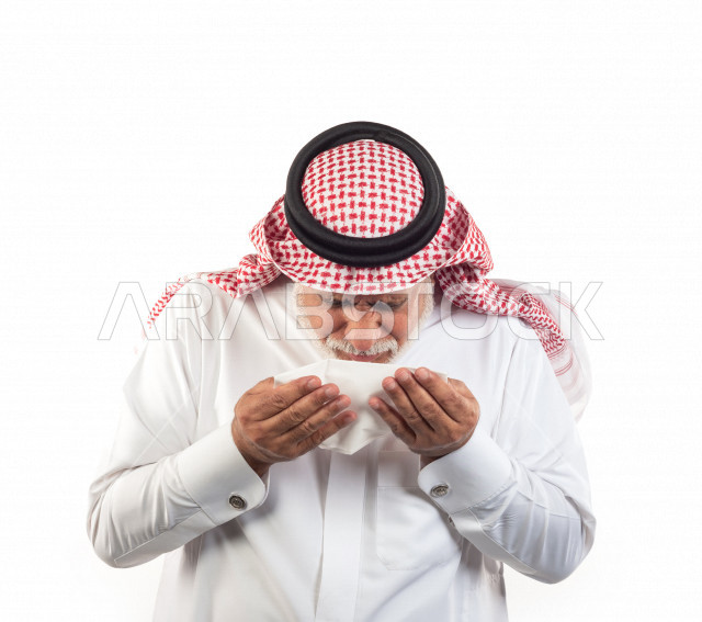 بورتريه رجل عربي سعودي كبير بالسن بيده منديل يضعه على أنفه والرغبة بالعطس , رجل شايب سعودي يرتدي ثوب أبيض مع شماغ وعقال , خلفية بيضاء , رجل مسن في حاله من العطس مفهوم لغة الجسد وتعابير الوجه