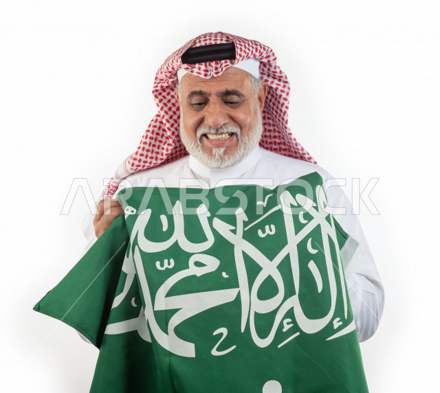 بورتريه رجل عربي سعودي كبير بالسن يحمل علم المملكة العربية السعودية وحالة من السعادة , يرتدي ثوب أبيض مع شماغ وعقال , خلفية بيضاء , رجل مسن متبسم وهو يحمل علم السعودية مفهوم تعبيرالوجه 