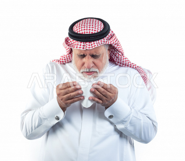 بورتريه رجل عربي سعودي كبير بالسن بيده منديل يضعه على أنفه والرغبة بالعطس , رجل شايب سعودي يرتدي ثوب أبيض مع شماغ وعقال , خلفية بيضاء , رجل مسن في حاله من العطس مفهوم لغة الجسد وتعابير الوجه