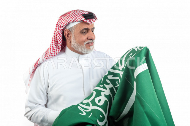 بورتريه رجل عربي سعودي كبير بالسن يحمل علم المملكة العربية السعودية وحالة من السعادة , يرتدي ثوب أبيض مع شماغ وعقال , خلفية بيضاء , رجل مسن متبسم وهو يحمل علم السعودية مفهوم تعبيرالوجه 