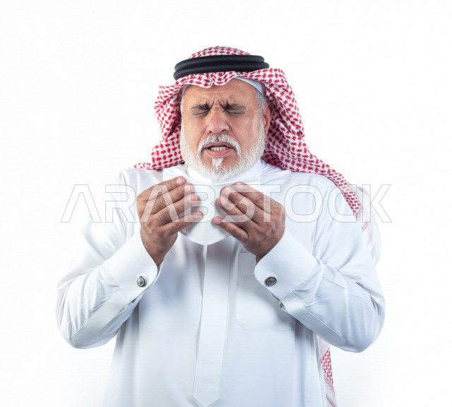 بورتريه رجل عربي سعودي كبير بالسن بيده منديل يضعه على أنفه والرغبة بالعطس , رجل شايب سعودي يرتدي ثوب أبيض مع شماغ وعقال , خلفية بيضاء , رجل مسن في حاله من العطس مفهوم لغة الجسد وتعابير الوجه