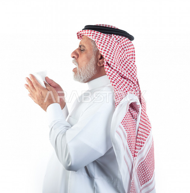 بورتريه رجل عربي سعودي كبير بالسن بيده منديل يضعه على أنفه والرغبة بالعطس , منظر جانبي لرجل شايب سعودي يرتدي ثوب أبيض مع شماغ وعقال , خلفية بيضاء , رجل مسن في حاله من العطس مفهوم لغة الجسد وتعابير الوجه