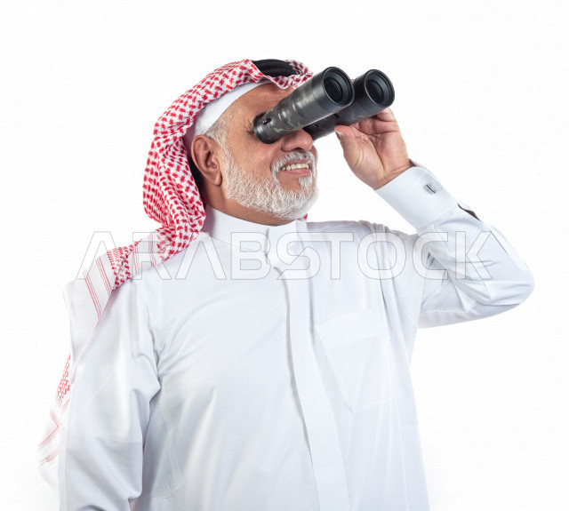 بورتريه رجل عربي سعودي كبير بالسن يقوم بالنظر من خلال المنظار الثنائي (التلسكوب) , صورة مقربة , شايب سعودي يرتدي ثوب أبيض مع شماغ وعقال , خلفية بيضاء , رجل مسن ينظر بالمنظار ثنائي العينين , مفهوم تعابيرالوجه ولغة الجسد
