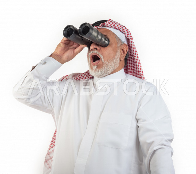 بورتريه رجل عربي سعودي كبير بالسن يقوم بالنظر من خلال المنظار الثنائي (التلسكوب) , صورة مقربة , شايب سعودي يرتدي ثوب أبيض مع شماغ وعقال , خلفية بيضاء , رجل مسن ينظر بالمنظار ثنائي العينين , مفهوم تعابيرالوجه ولغة الجسد