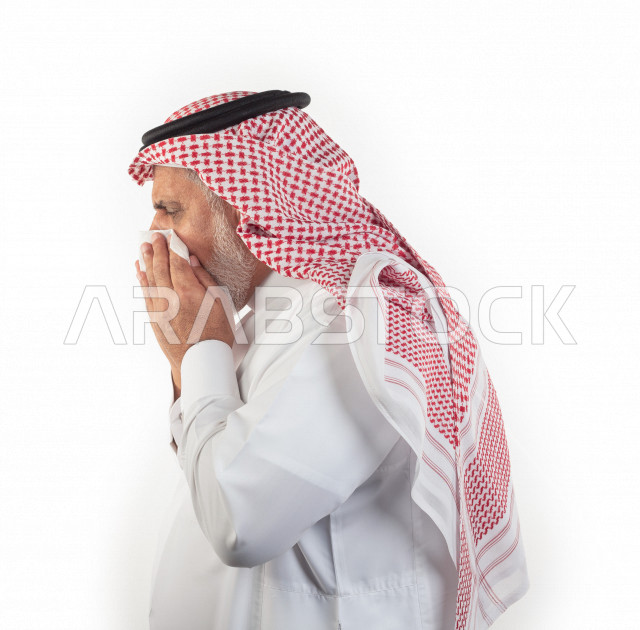 بورتريه رجل عربي سعودي كبير بالسن بيده منديل يضعه على أنفه والرغبة بالعطس , منظر جانبي لرجل شايب سعودي يرتدي ثوب أبيض مع شماغ وعقال , خلفية بيضاء , رجل مسن في حاله من العطس مفهوم لغة الجسد وتعابير الوجه