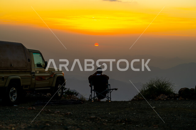 The distinctive landscapes of the Kingdom of Saudi Arabia, a silhouette from behind of a man sitting on top of the Al Hada Mountains watching the sunset, recreational trips in the famous tourist areas in the city of Taif