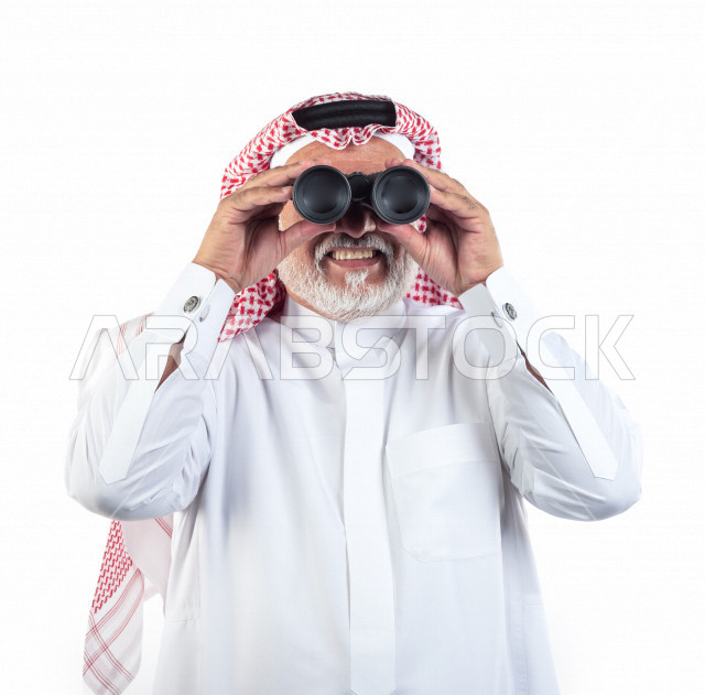 بورتريه رجل عربي سعودي كبير بالسن يقوم بالنظر من خلال المنظار الثنائي (التلسكوب) , صورة مقربة , شايب سعودي يرتدي ثوب أبيض مع شماغ وعقال , خلفية بيضاء , رجل مسن ينظر بالمنظار ثنائي العينين , مفهوم تعابيرالوجه ولغة الجسد
