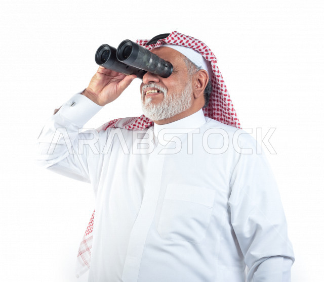 بورتريه رجل عربي سعودي كبير بالسن يقوم بالنظر من خلال المنظار الثنائي (التلسكوب) , صورة مقربة , شايب سعودي يرتدي ثوب أبيض مع شماغ وعقال , خلفية بيضاء , رجل مسن ينظر بالمنظار ثنائي العينين , مفهوم تعابيرالوجه ولغة الجسد