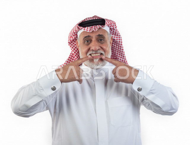 بورتريه رجل عربي سعودي كبير بالسن يشير بيده على أسنانه , صورة مقربة , شايب سعودي يرتدي ثوب أبيض مع شماغ وعقال , خلفية بيضاء , رجل مسن يشير أصبعه السبابه على أسنانه , مفهوم تعابيرالوجه ولغة الجسد