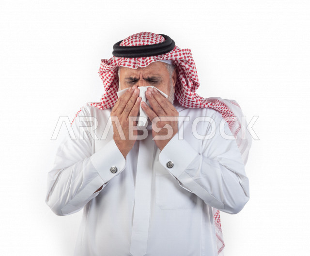 بورتريه رجل عربي سعودي كبير بالسن بيده منديل يضعه على أنفه والرغبة بالعطس , رجل شايب سعودي يرتدي ثوب أبيض مع شماغ وعقال , خلفية بيضاء , رجل مسن في حاله من العطس مفهوم لغة الجسد وتعابير الوجه