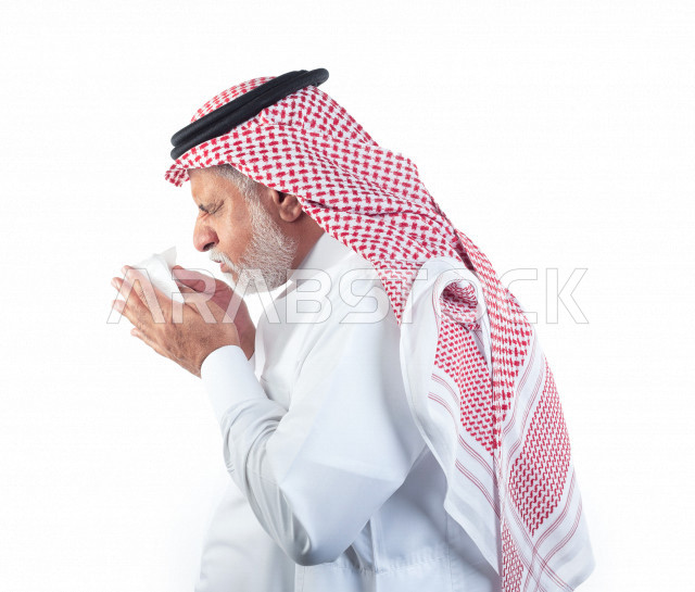 بورتريه رجل عربي سعودي كبير بالسن بيده منديل يضعه على أنفه والرغبة بالعطس , منظر جانبي لرجل شايب سعودي يرتدي ثوب أبيض مع شماغ وعقال , خلفية بيضاء , رجل مسن في حاله من العطس مفهوم لغة الجسد وتعابير الوجه