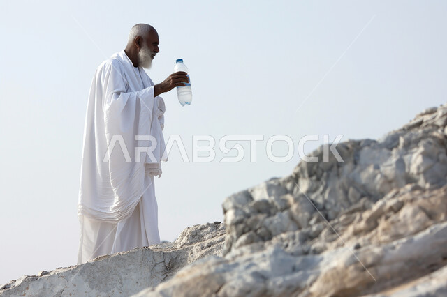 مفهوم العبادة والتقرب الى الله، أداء مناسك الحج والعمرة، رجل كبير السن عربي سعودي خليجي يرتدي ملابس الاحرام يمسك بيده عبوة الماء يقف على جبل عرفات (جبل الرحمة)، أماكن ومعالم دينية اسلامية مقدسة في المملكة العربية السعودية