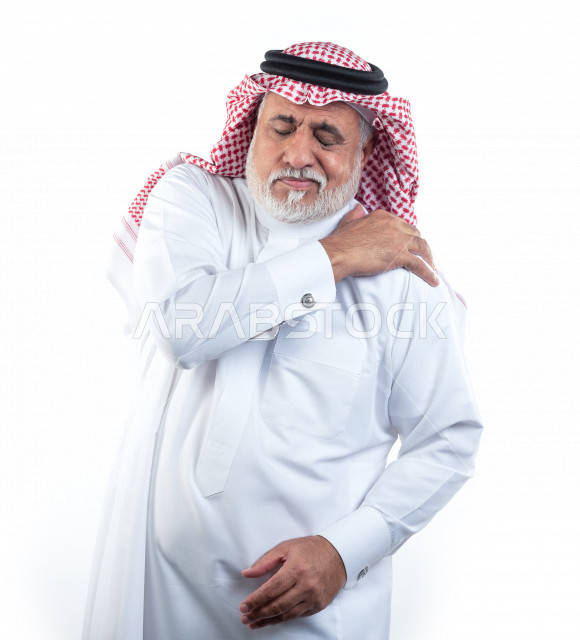 بورتريه رجل عربي سعودي كبير بالسن يضع يده على كتفه يشعر بحالة من الوجع والألم , صورة مقربة , شايب سعودي يرتدي ثوب أبيض مع شماغ وعقال , خلفية بيضاء , رجل مسن يشعر بالتعب والألم الشديد في كتف يده اليمين , مفهوم تعابيرالوجه ولغة الجسد
