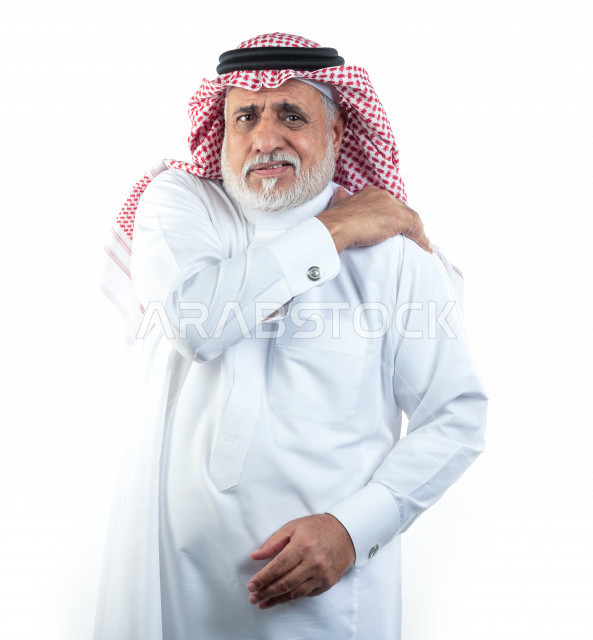 بورتريه رجل عربي سعودي كبير بالسن يضع يده على كتفه يشعر بحالة من الوجع والألم , صورة مقربة , شايب سعودي يرتدي ثوب أبيض مع شماغ وعقال , خلفية بيضاء , رجل مسن يشعر بالتعب والألم الشديد في كتف يده اليمين , مفهوم تعابيرالوجه ولغة الجسد