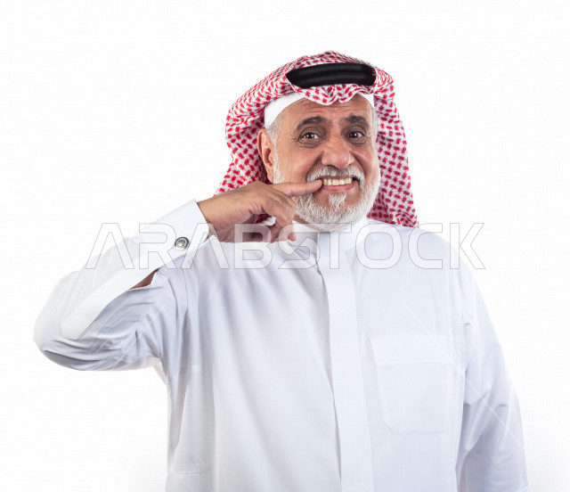 بورتريه رجل عربي سعودي كبير بالسن يشير بيده على أسنانه , صورة مقربة , شايب سعودي يرتدي ثوب أبيض مع شماغ وعقال , خلفية بيضاء , رجل مسن يشير أصبعه السبابه على أسنانه , مفهوم تعابيرالوجه ولغة الجسد