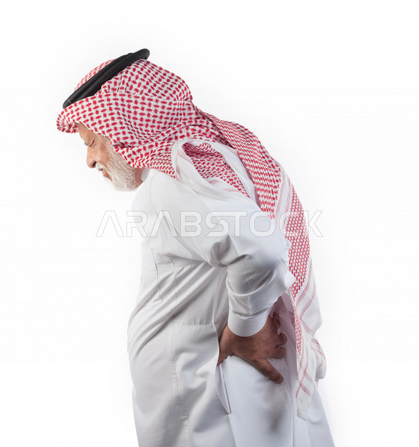 بورتريه رجل عربي سعودي كبير بالسن يضع يده على ظهره يشعر بحالة من الوجع والألم , صورة مقربة , شايب سعودي يرتدي ثوب أبيض مع شماغ وعقال , خلفية بيضاء , منظر جانبي لرجل مسن يشعر بالتعب والألم الشديد في ظهره , مفهوم تعابيرالوجه ولغة الجسد