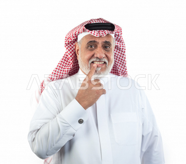 بورتريه رجل عربي سعودي كبير بالسن يشير بيده على أسنانه , صورة مقربة , شايب سعودي يرتدي ثوب أبيض مع شماغ وعقال , خلفية بيضاء , رجل مسن يشير أصبعه السبابه على أسنانه , مفهوم تعابيرالوجه ولغة الجسد