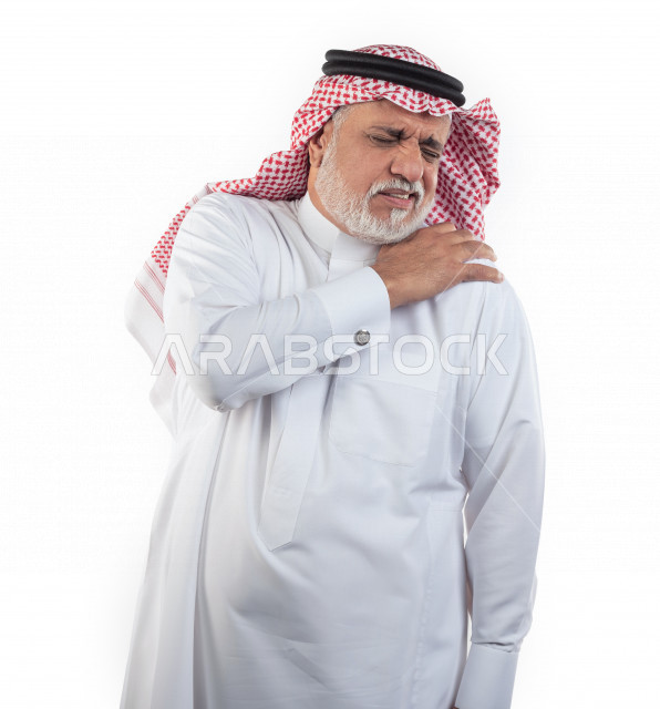 بورتريه رجل عربي سعودي كبير بالسن يضع يده على كتفه يشعر بحالة من الوجع والألم , صورة مقربة , شايب سعودي يرتدي ثوب أبيض مع شماغ وعقال , خلفية بيضاء , رجل مسن يشعر بالتعب والألم الشديد في كتف يده اليمين , مفهوم تعابيرالوجه ولغة الجسد