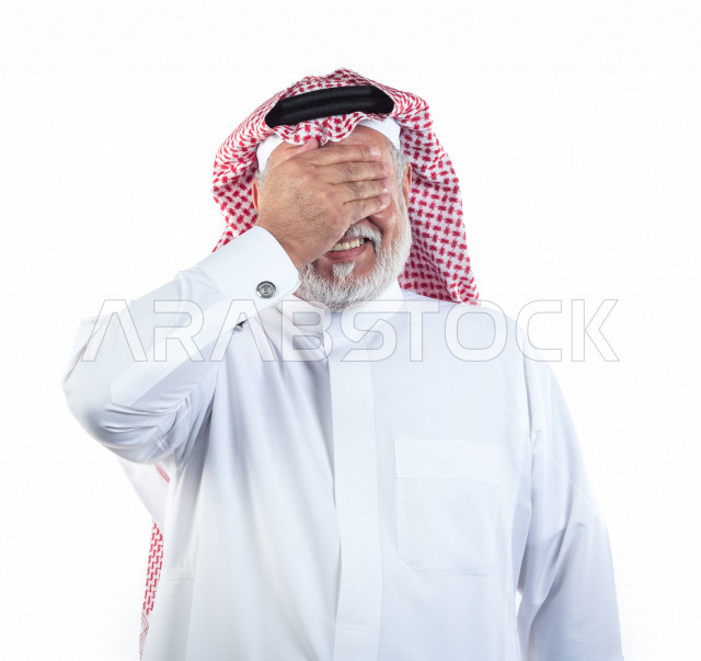 بورتريه رجل عربي سعودي كبير بالسن يضع يده على عينيه بإيماءات وجه ويدين مختلفة , صورة مقربة , شايب سعودي يرتدي ثوب أبيض مع شماغ وعقال , خلفية بيضاء , رجل مسن يلتقط الصور وهو مبتسم ويده على عينيه , مفهوم تعابيرالوجه ولغة الجسد المختلفة