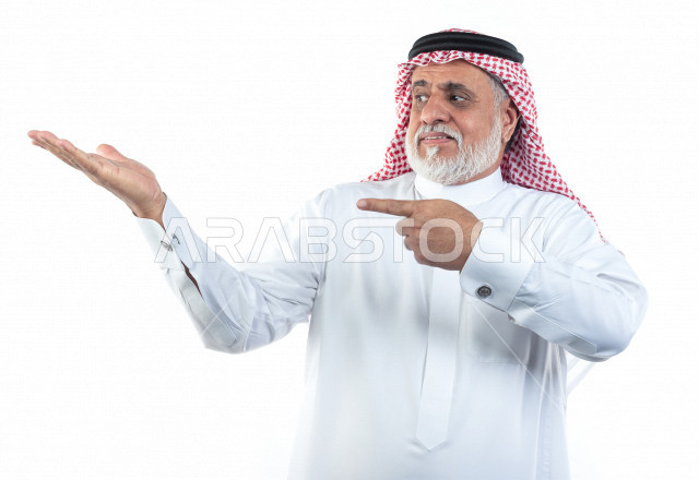 بورتريه رجل عربي سعودي كبير بالسن يشير بإصبعه على كف يده وهو يبتسم بإيماءات وجه ويدين مختلفة , صورة مقربة , شايب سعودي يرتدي ثوب أبيض مع شماغ وعقال , خلفية بيضاء , رجل مسن يلتقط الصور ويشير أصبعه على يده , مفهوم تعابيرالوجه ولغة الجسد المختلفة