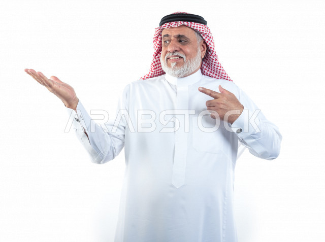 بورتريه رجل عربي سعودي كبير بالسن يشير بإصبعه على كف يده وهو يبتسم بإيماءات وجه ويدين مختلفة , صورة مقربة , شايب سعودي يرتدي ثوب أبيض مع شماغ وعقال , خلفية بيضاء , رجل مسن يلتقط الصور ويشير أصبعه على يده , مفهوم تعابيرالوجه ولغة الجسد المختلفة